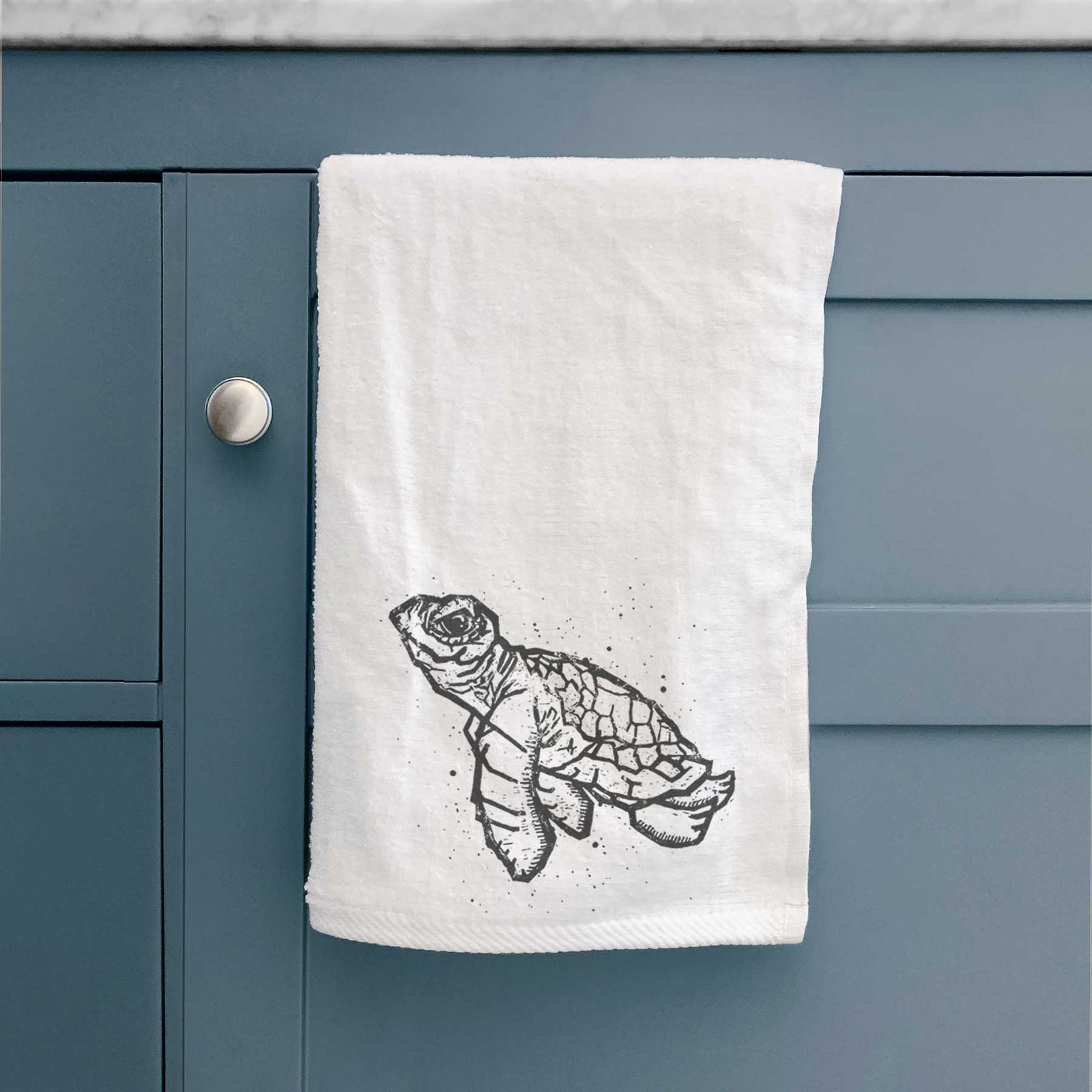 White hand towel printed with a black linocut-style illustration of a baby sea turtle with a patterned shell and ink splatters.