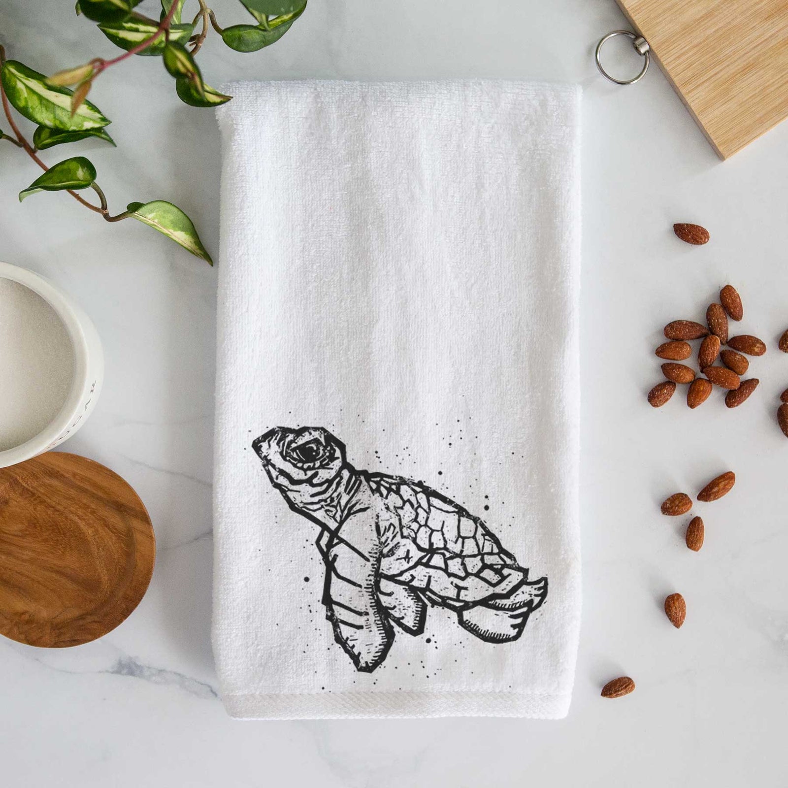 White hand towel printed with a black linocut-style illustration of a baby sea turtle with a patterned shell and ink splatters.