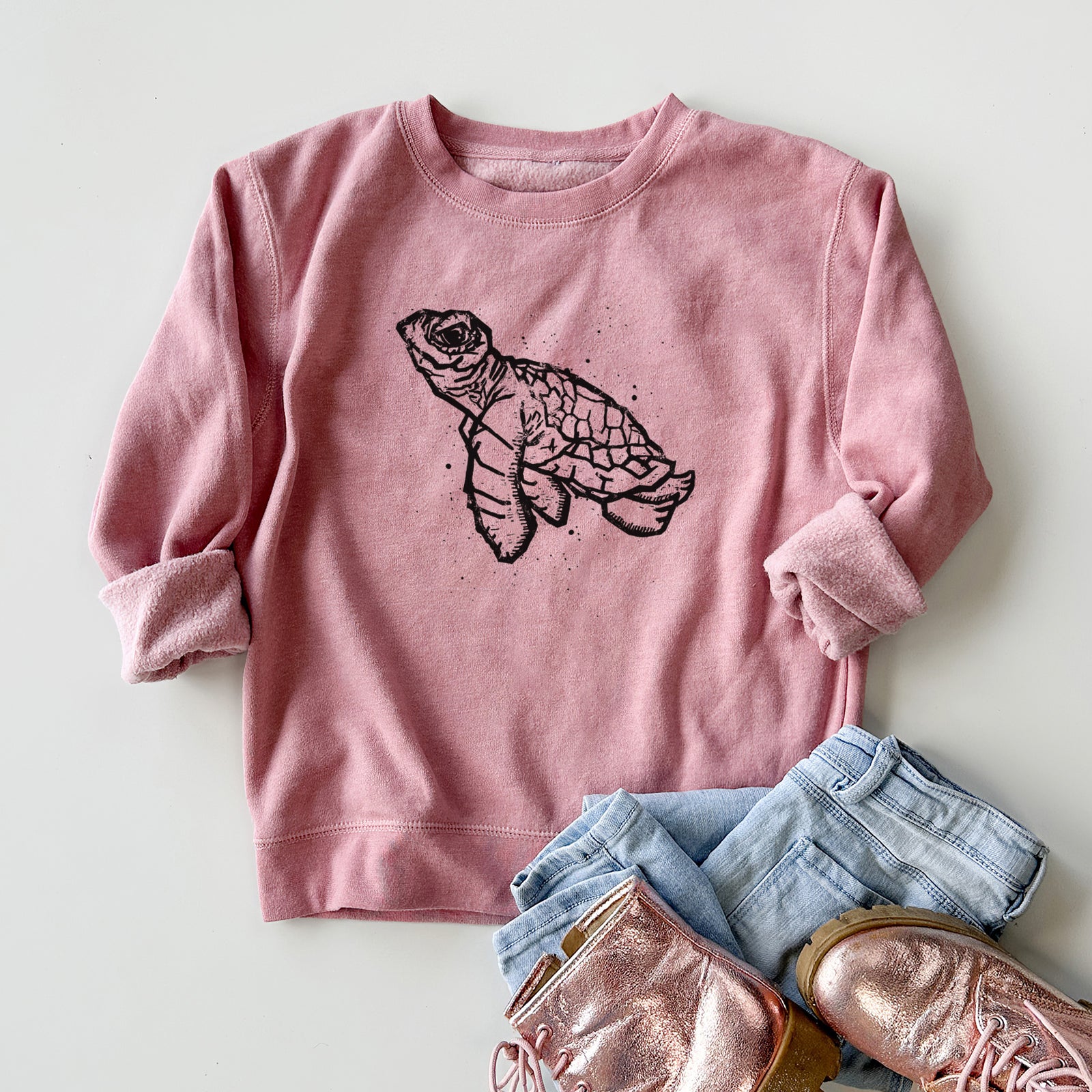 Boy wearing a youth crewneck sweatshirt printed with a black linocut-style illustration of a baby sea turtle with a patterned shell and ink splatters, outdoors.