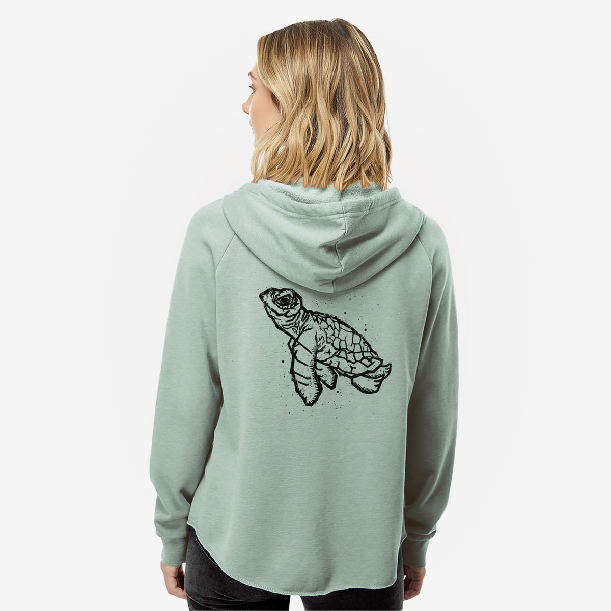Woman wearing a sage green women's Cali Wave zip-up sweatshirt printed with a black linocut-style illustration of a baby sea turtle with a patterned shell and ink splatters, seen from behind.
