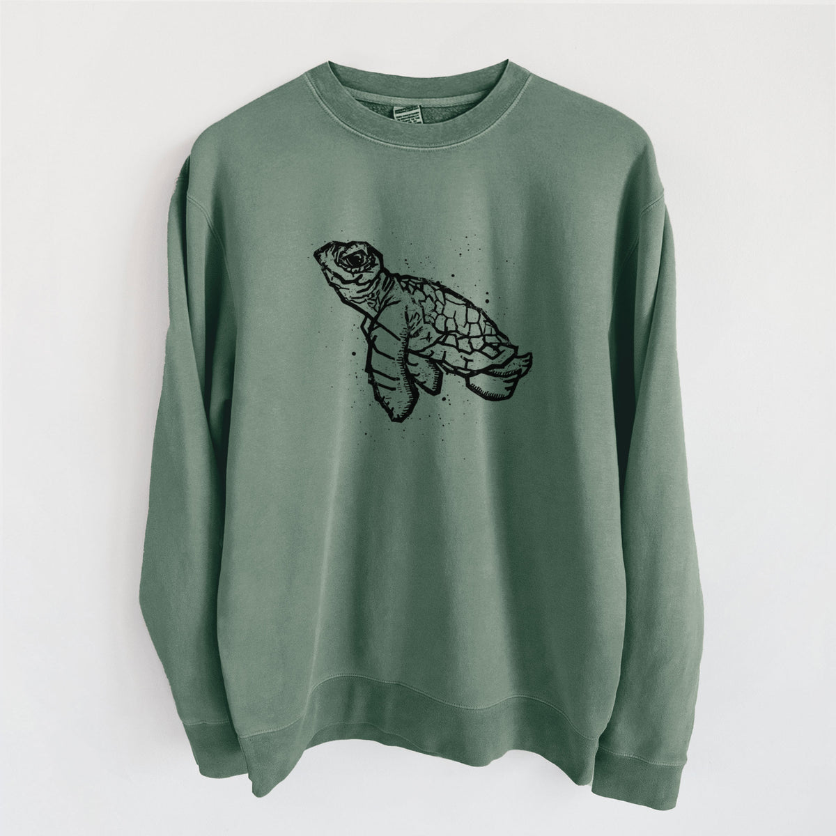 Alpine green pigment dyed crewneck sweatshirt laid flat, printed with a black linocut-style illustration of a baby sea turtle with a patterned shell and ink splatters.