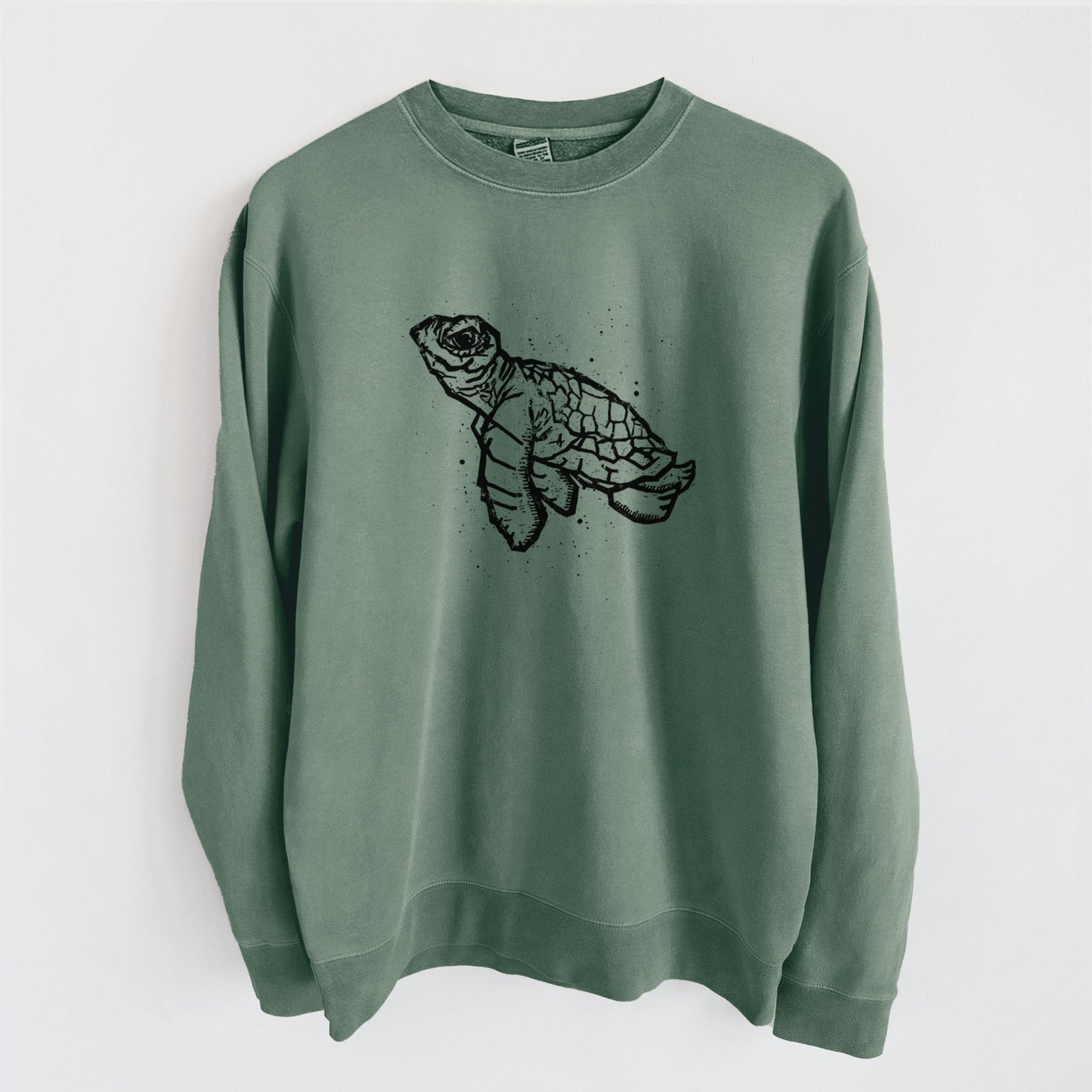 Alpine green pigment dyed crewneck sweatshirt laid flat, printed with a black linocut-style illustration of a baby sea turtle with a patterned shell and ink splatters.