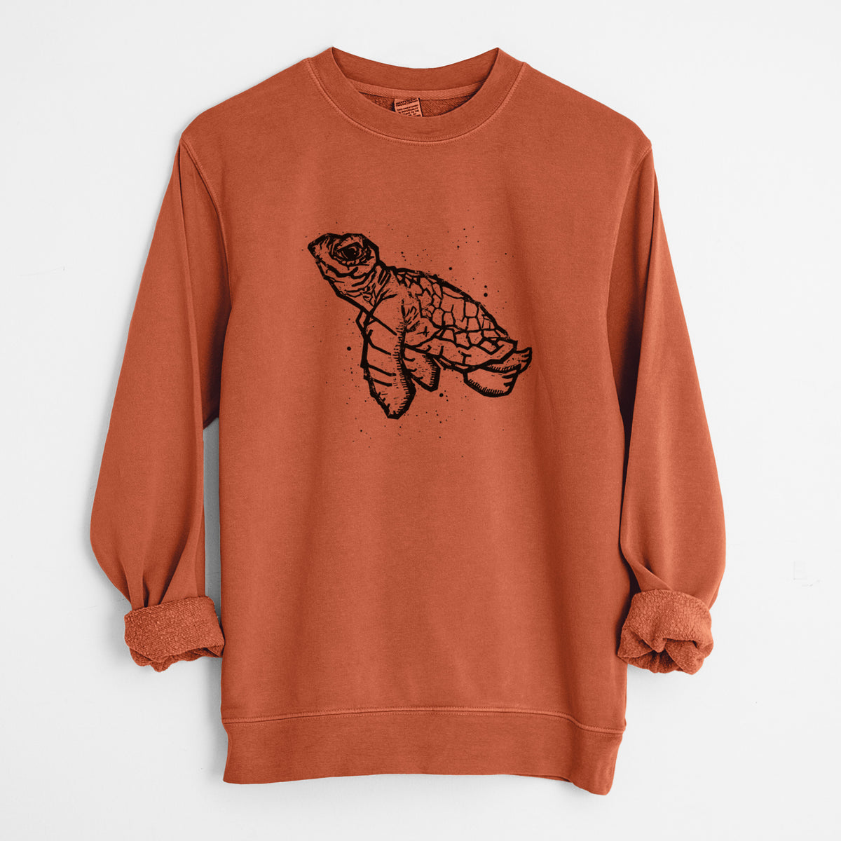 Amber orange pigment dyed crewneck sweatshirt laid flat with the sleeves rolled, printed with a black linocut-style illustration of a baby sea turtle with a patterned shell and ink splatters.