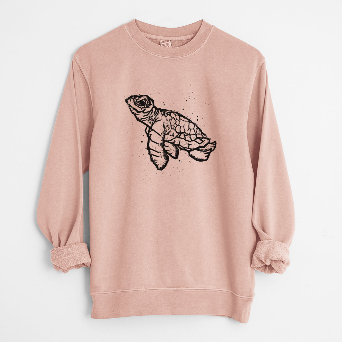 Dusty pink pigment dyed crewneck sweatshirt laid flat with the sleeves rolled, printed with a black linocut-style illustration of a baby sea turtle with a patterned shell and ink splatters.
