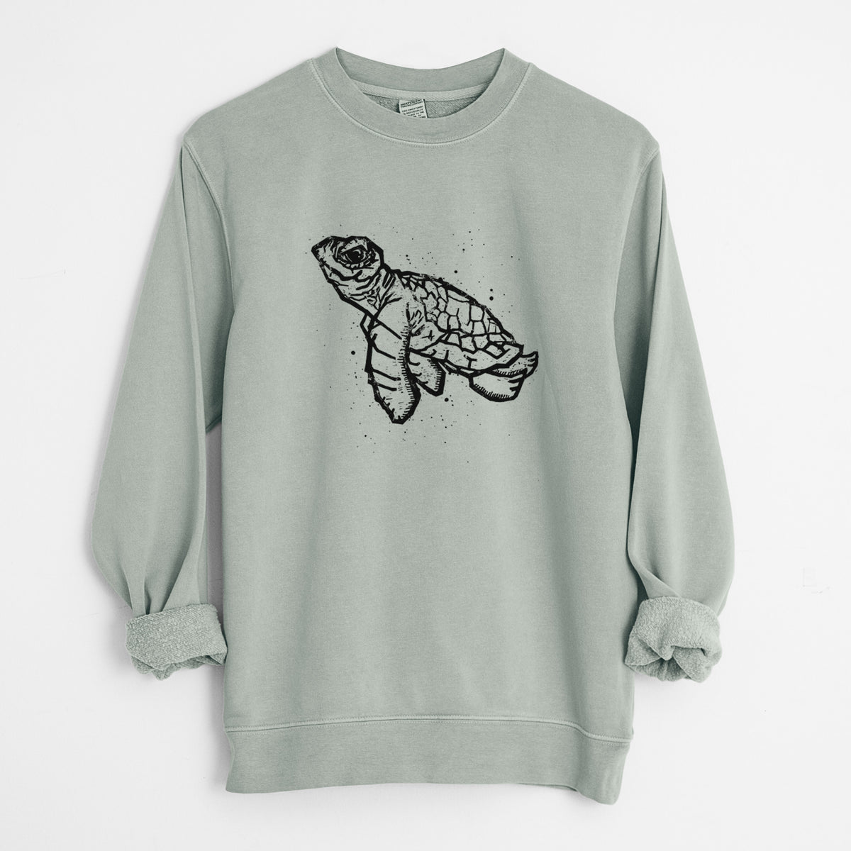 Sage green pigment dyed crewneck sweatshirt laid flat with the sleeves rolled, printed with a black linocut-style illustration of a baby sea turtle with a patterned shell and ink splatters.