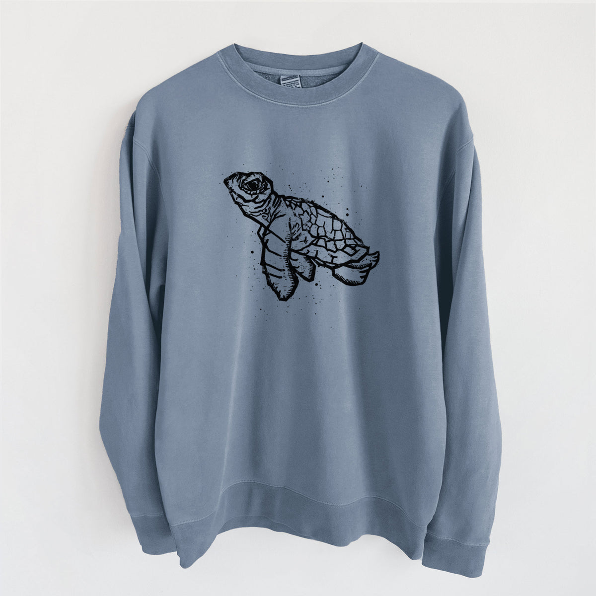Slate blue pigment dyed crewneck sweatshirt laid flat, printed with a black linocut-style illustration of a baby sea turtle with a patterned shell and ink splatters.