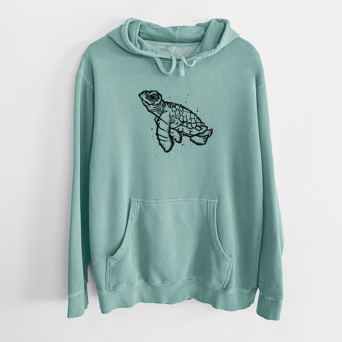 Mint green pigment dyed hoodie laid flat, printed with a black linocut-style illustration of a baby sea turtle with a patterned shell and ink splatters.