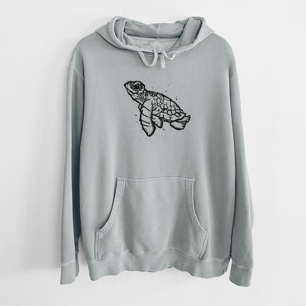Sage green pigment dyed hoodie laid flat, printed with a black linocut-style illustration of a baby sea turtle with a patterned shell and ink splatters.
