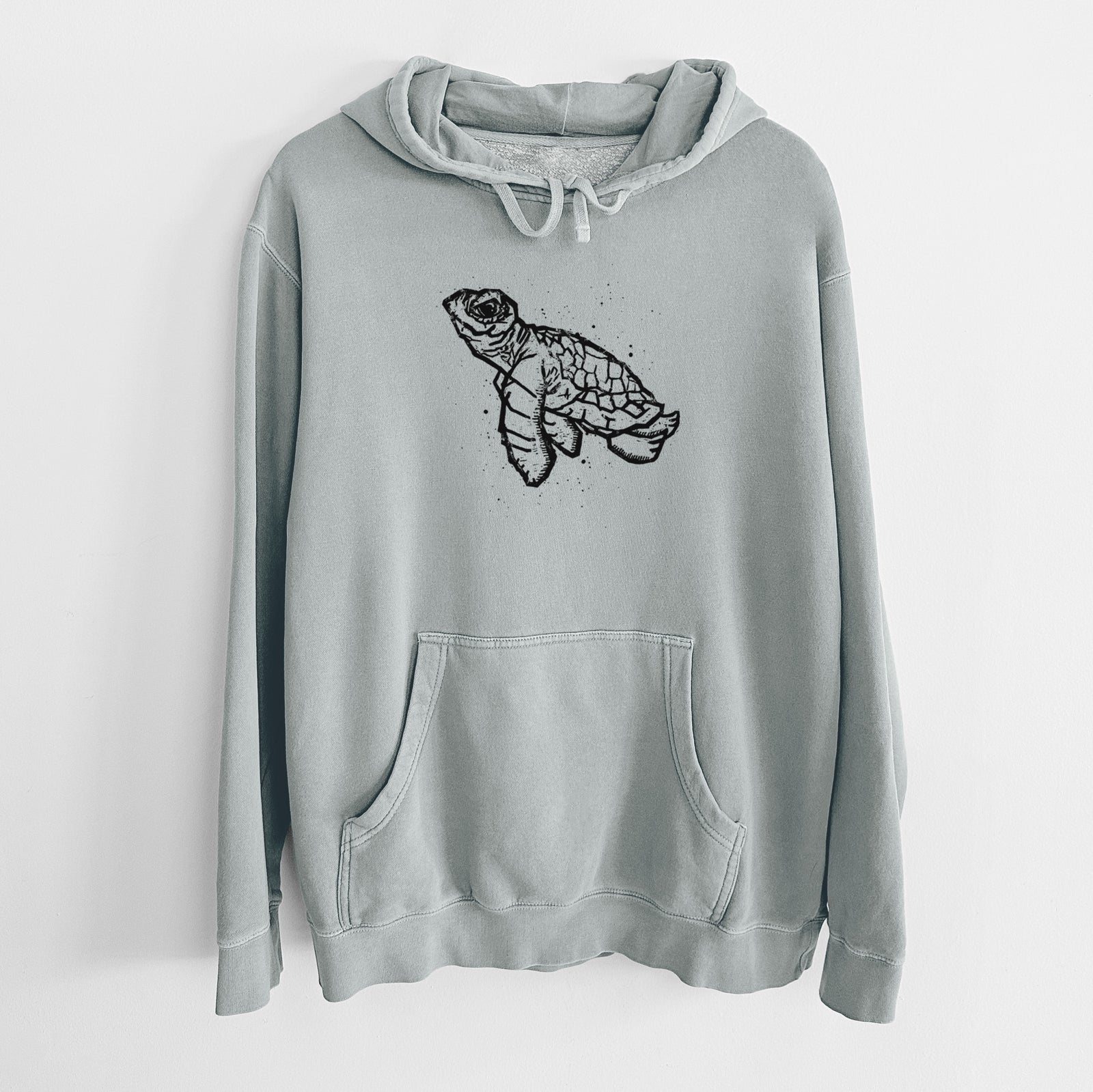 Sage green pigment dyed hoodie laid flat, printed with a black linocut-style illustration of a baby sea turtle with a patterned shell and ink splatters.