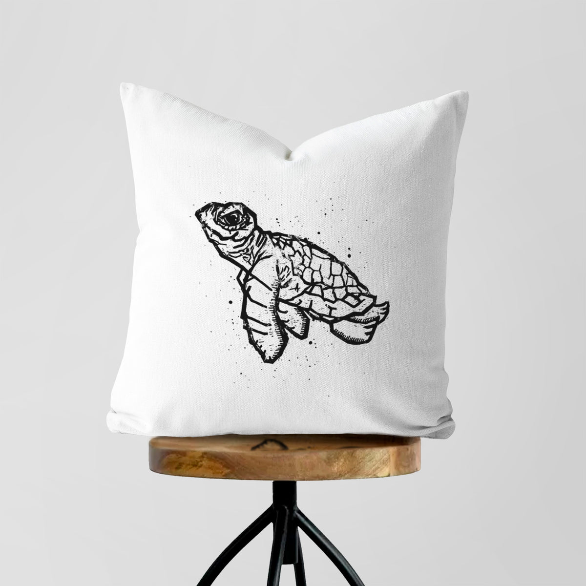 White pillow cover printed with a black linocut-style illustration of a baby sea turtle with a patterned shell and ink splatters, on a wooden stool.