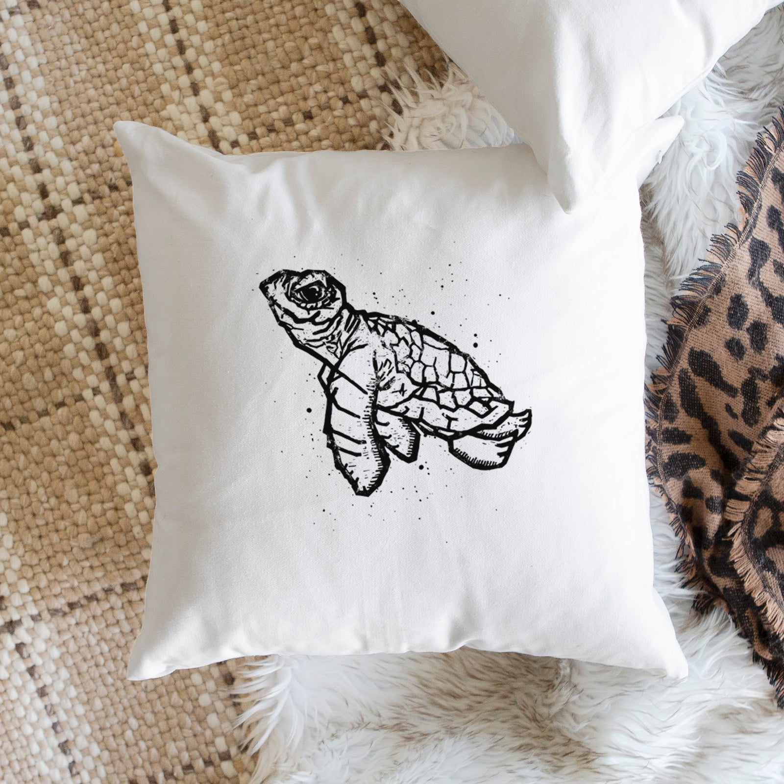 White pillow cover printed with a black linocut-style illustration of a baby sea turtle with a patterned shell and ink splatters, on a bench beside plants.
