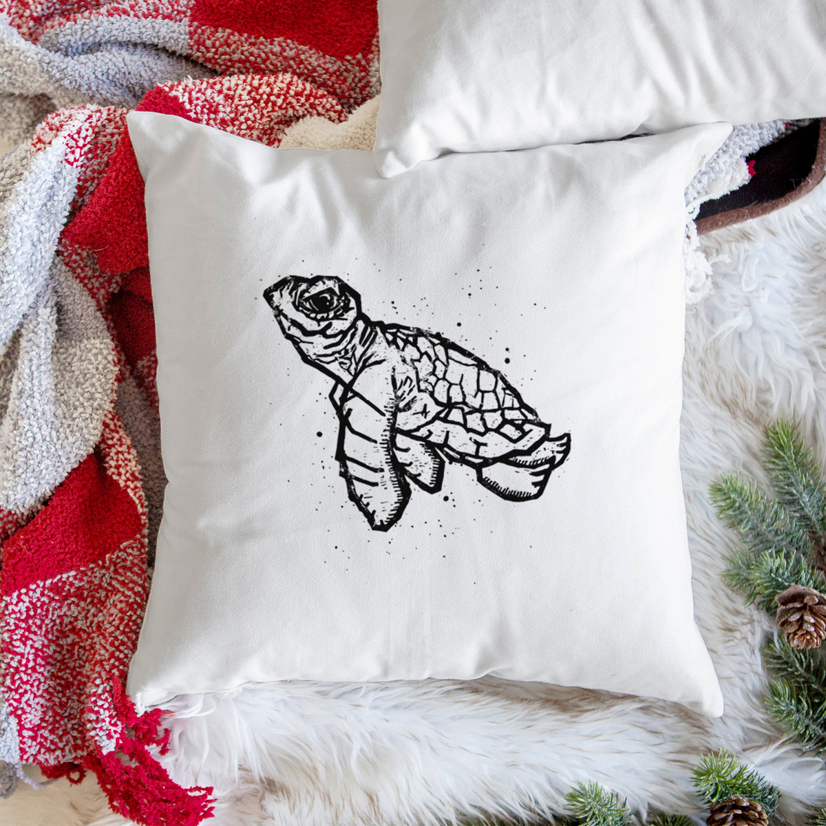 White pillow cover printed with a black linocut-style illustration of a baby sea turtle with a patterned shell and ink splatters, on a woven chair.