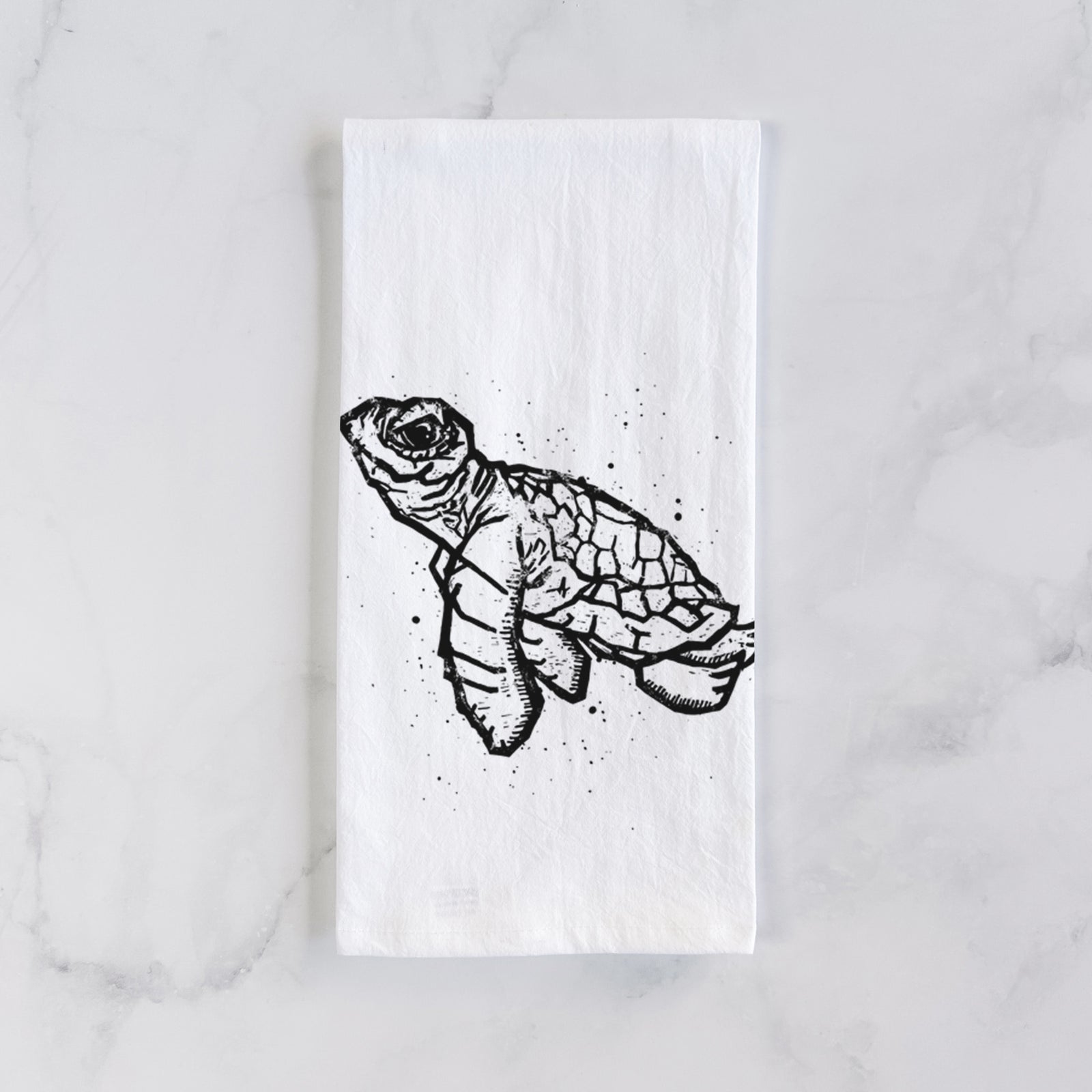 White cotton tea towel printed with a black linocut-style illustration of a baby sea turtle with a patterned shell and ink splatters, on a marble counter beside a round wooden cutting board and greenery.