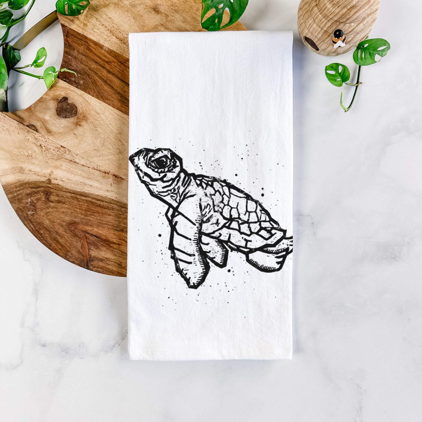 White cotton tea towel printed with a black linocut-style illustration of a baby sea turtle with a patterned shell and ink splatters, on a marble counter beside a round wooden cutting board and greenery.