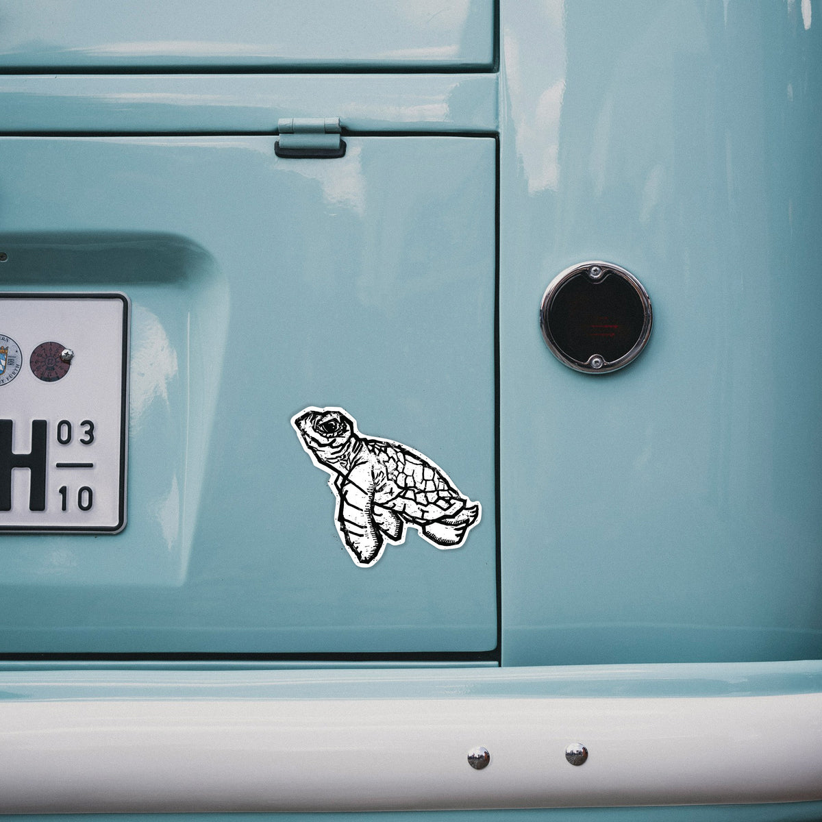 4 inch vinyl decal of a black linocut-style illustration of a baby sea turtle with a patterned shell and ink splatters on the back of a van.