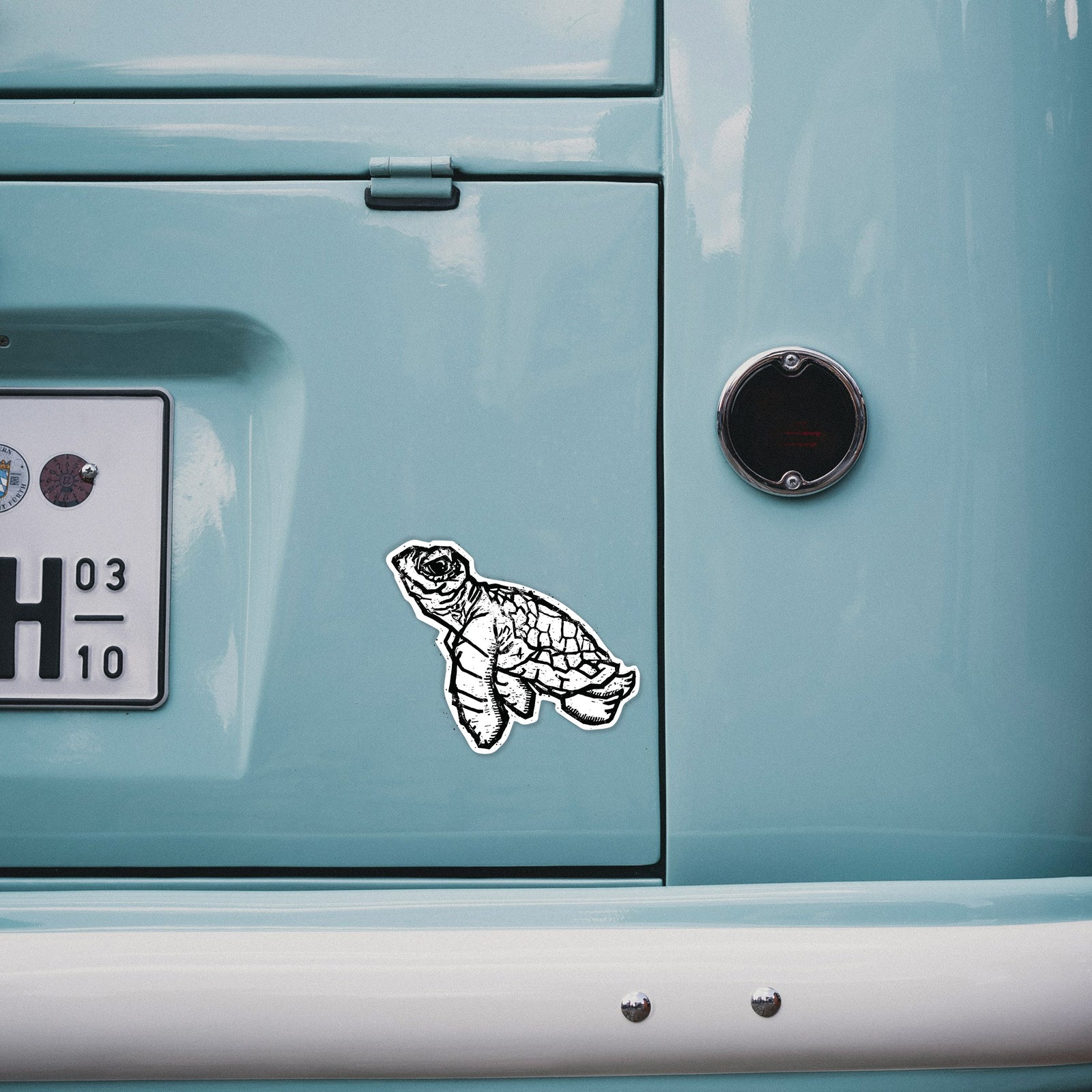 Hand holding a 4 inch vinyl decal of a black linocut-style illustration of a baby sea turtle with a patterned shell and ink splatters.