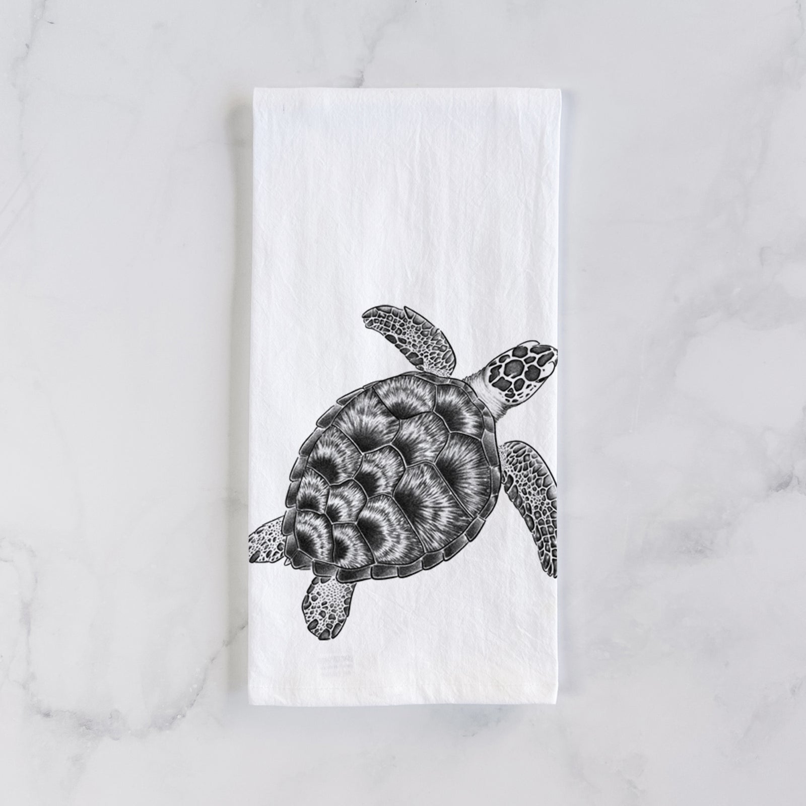 White cotton tea towel printed with a detailed black ink illustration of a loggerhead sea turtle swimming, seen from above, on a marble counter beside a round wooden cutting board and greenery.