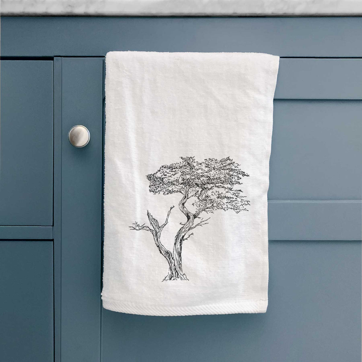 White hand towel printed with a black ink illustration of the windswept Lone Cypress tree on a rocky outcrop, hanging on a cabinet door.