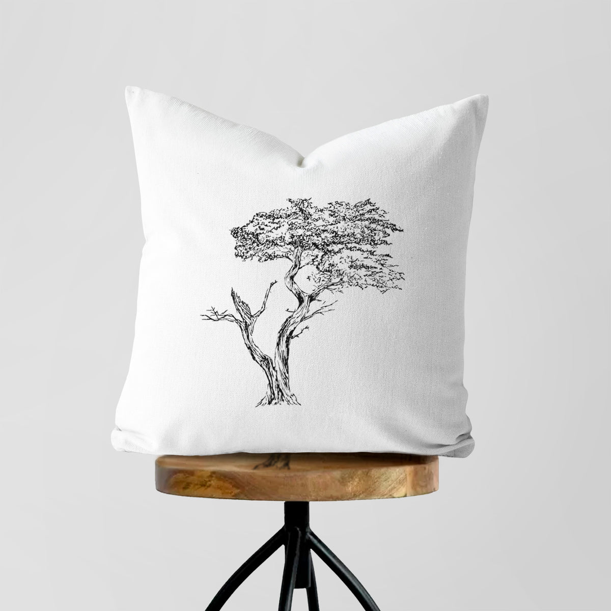 White pillow cover printed with a black ink illustration of the windswept Lone Cypress tree on a rocky outcrop, on a wooden stool.