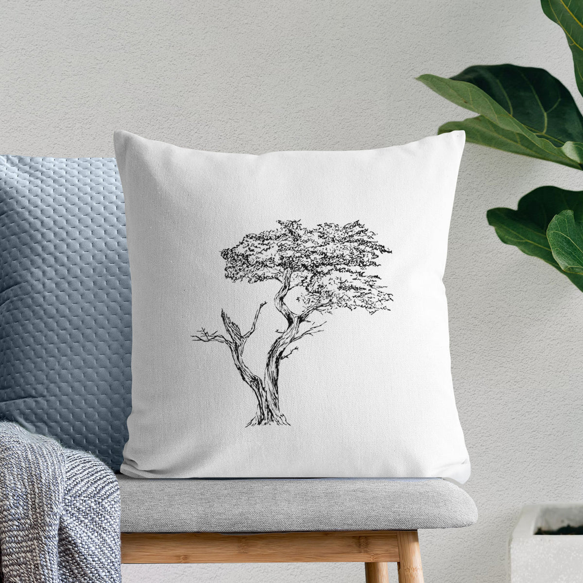 White pillow cover printed with a black ink illustration of the windswept Lone Cypress tree on a rocky outcrop, on a bench beside plants.