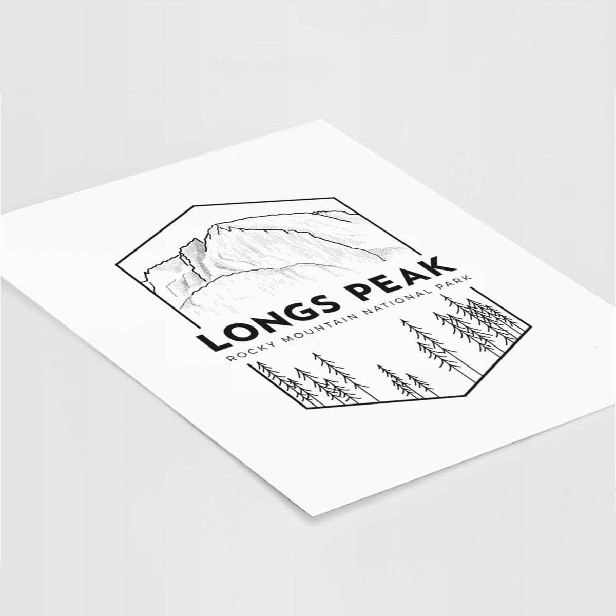 Art print of a black line art badge of Longs Peak above a pine forest, with the words Longs Peak, Rocky Mountain National Park, lying at an angle on a white surface.