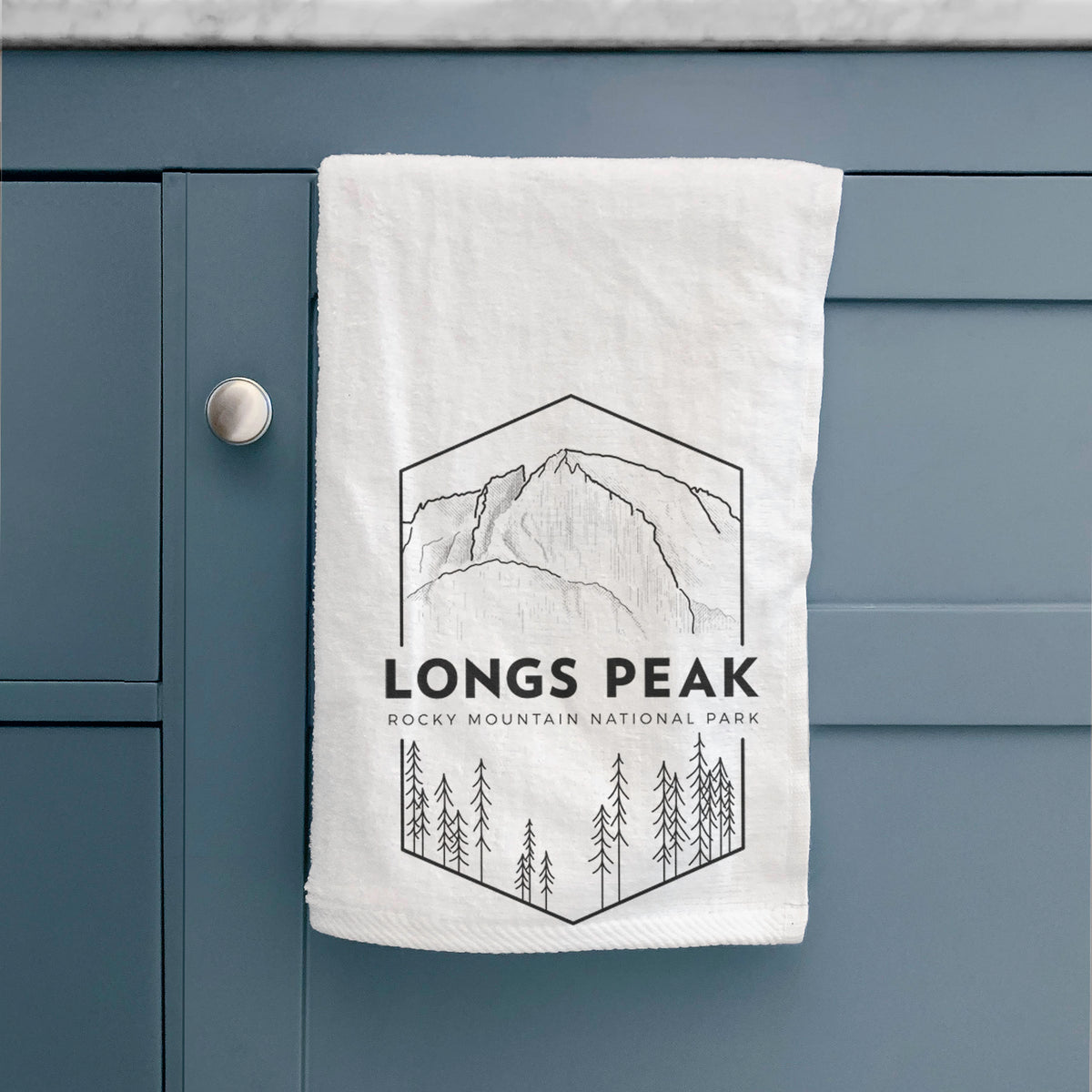 White hand towel printed with a black line art badge of Longs Peak above a pine forest, with the words Longs Peak, Rocky Mountain National Park, hanging on a cabinet door.