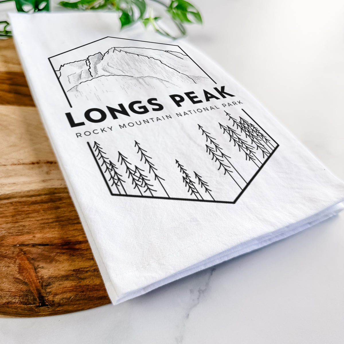 Close-up of a folded white tea towel on a wooden board, showing a black line art badge of Longs Peak above a pine forest, with the words Longs Peak, Rocky Mountain National Park.
