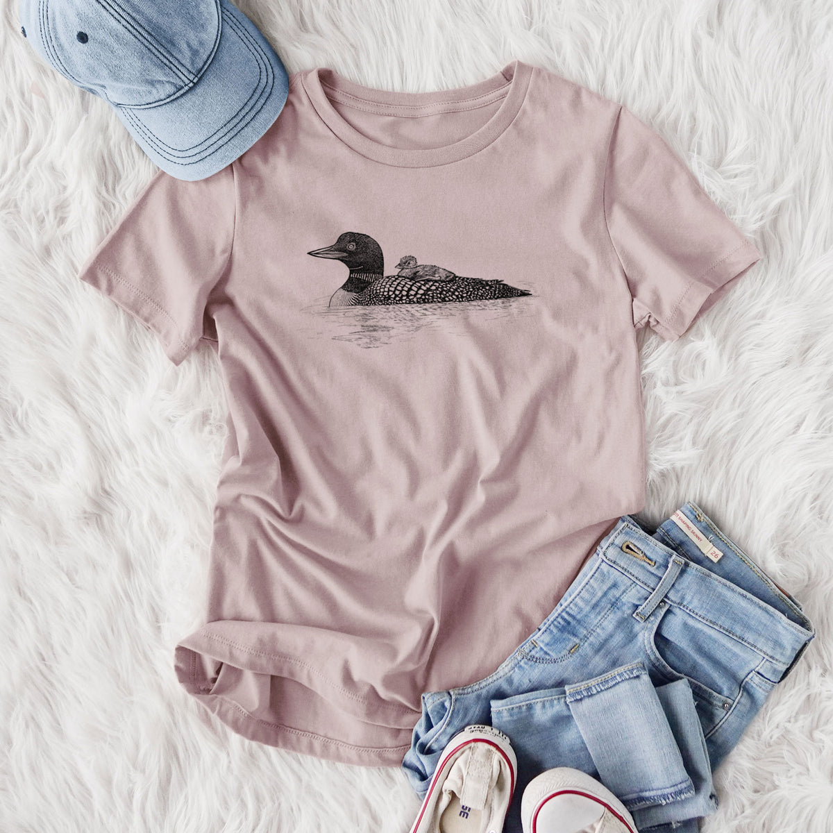 Women's relaxed fit cotton t-shirt printed with a black ink sketch of a common loon floating on water with a chick riding on its back, styled flat on a white faux fur rug with jeans.