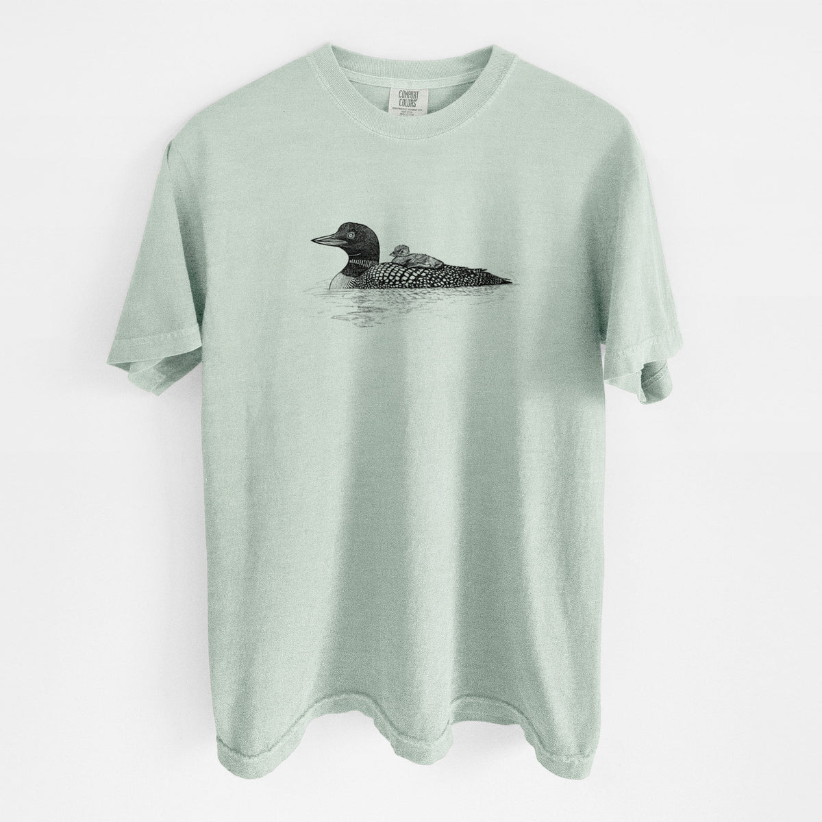 Bay green Comfort Colors t-shirt laid flat, printed with a black ink sketch of a common loon floating on water with a chick riding on its back.