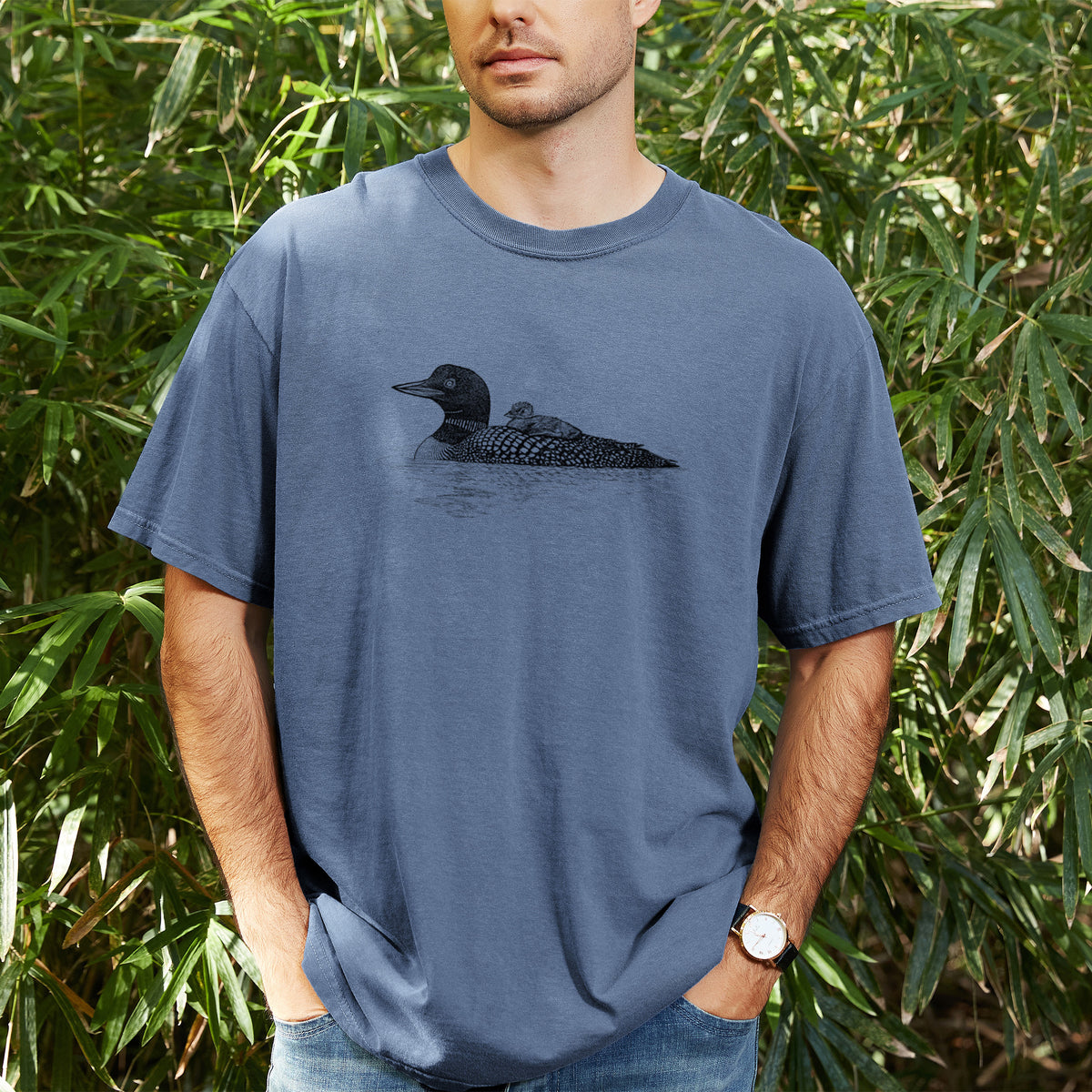Man wearing a Comfort Colors t-shirt printed with a black ink sketch of a common loon floating on water with a chick riding on its back, outdoors.