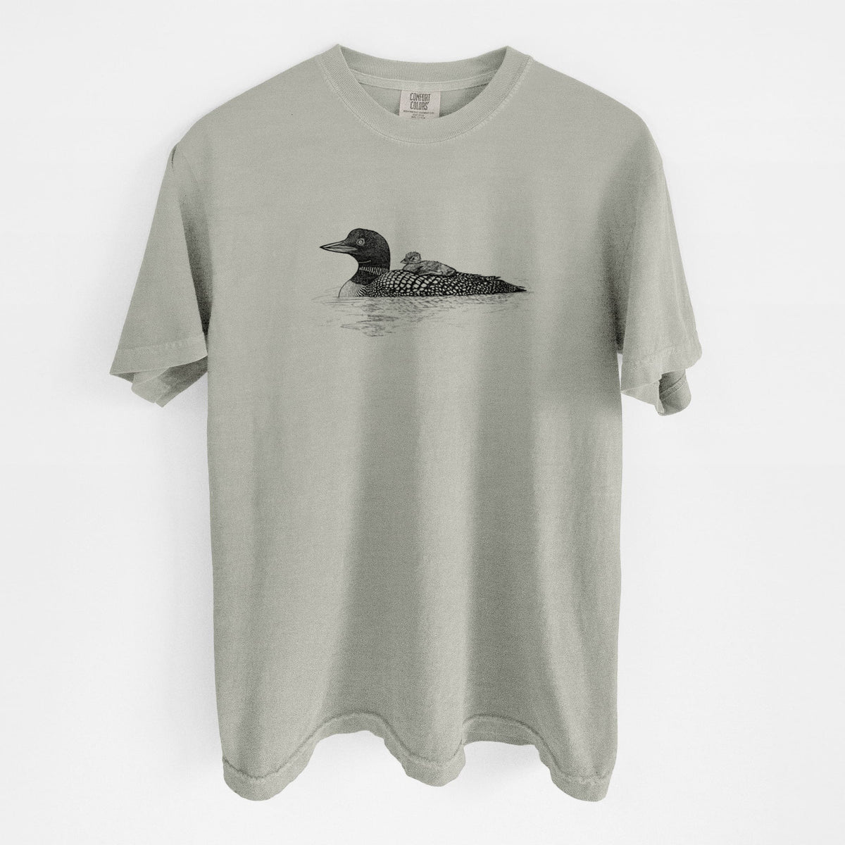 Sandstone beige Comfort Colors t-shirt laid flat, printed with a black ink sketch of a common loon floating on water with a chick riding on its back.