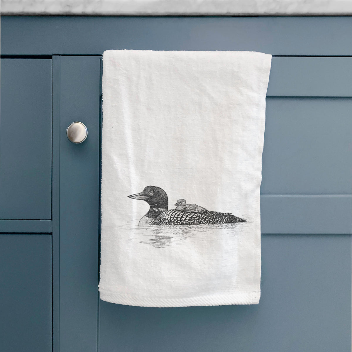 White hand towel printed with a black ink sketch of a common loon floating on water with a chick riding on its back, hanging on a cabinet door.