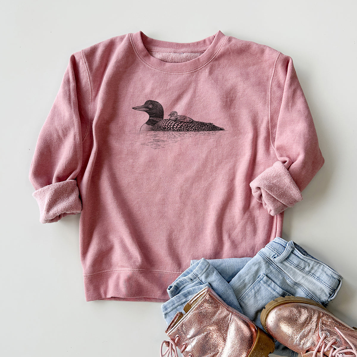Youth crewneck sweatshirt printed with a black ink sketch of a common loon floating on water with a chick riding on its back, styled flat with jeans and boots.
