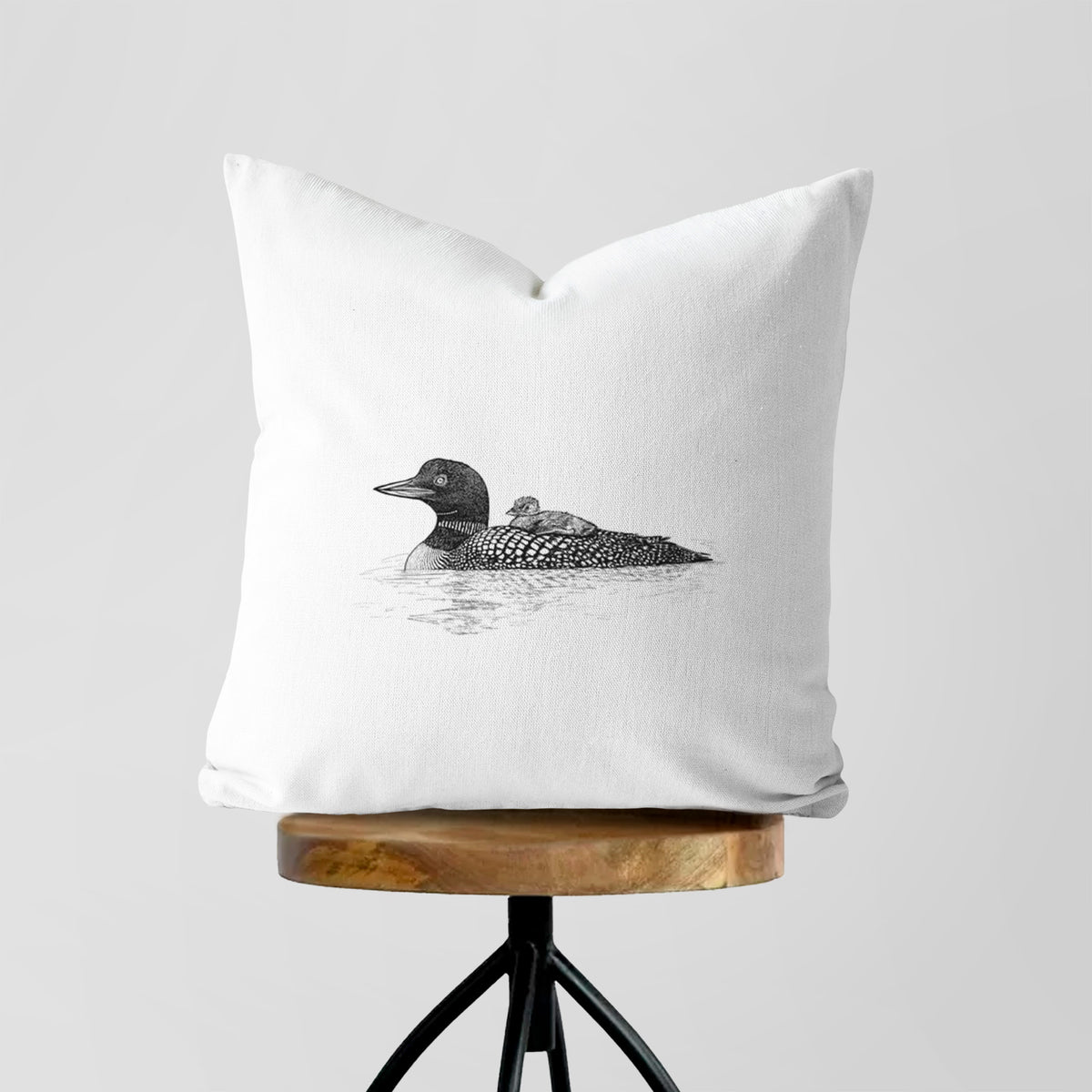 White pillow cover printed with a black ink sketch of a common loon floating on water with a chick riding on its back, on a wooden stool.