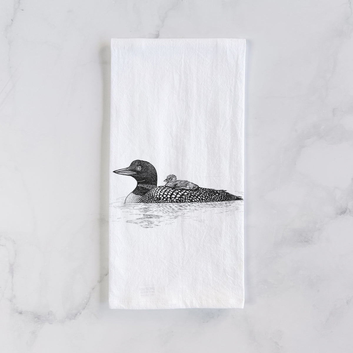 White cotton tea towel laid flat on marble, printed with a black ink sketch of a common loon floating on water with a chick riding on its back.