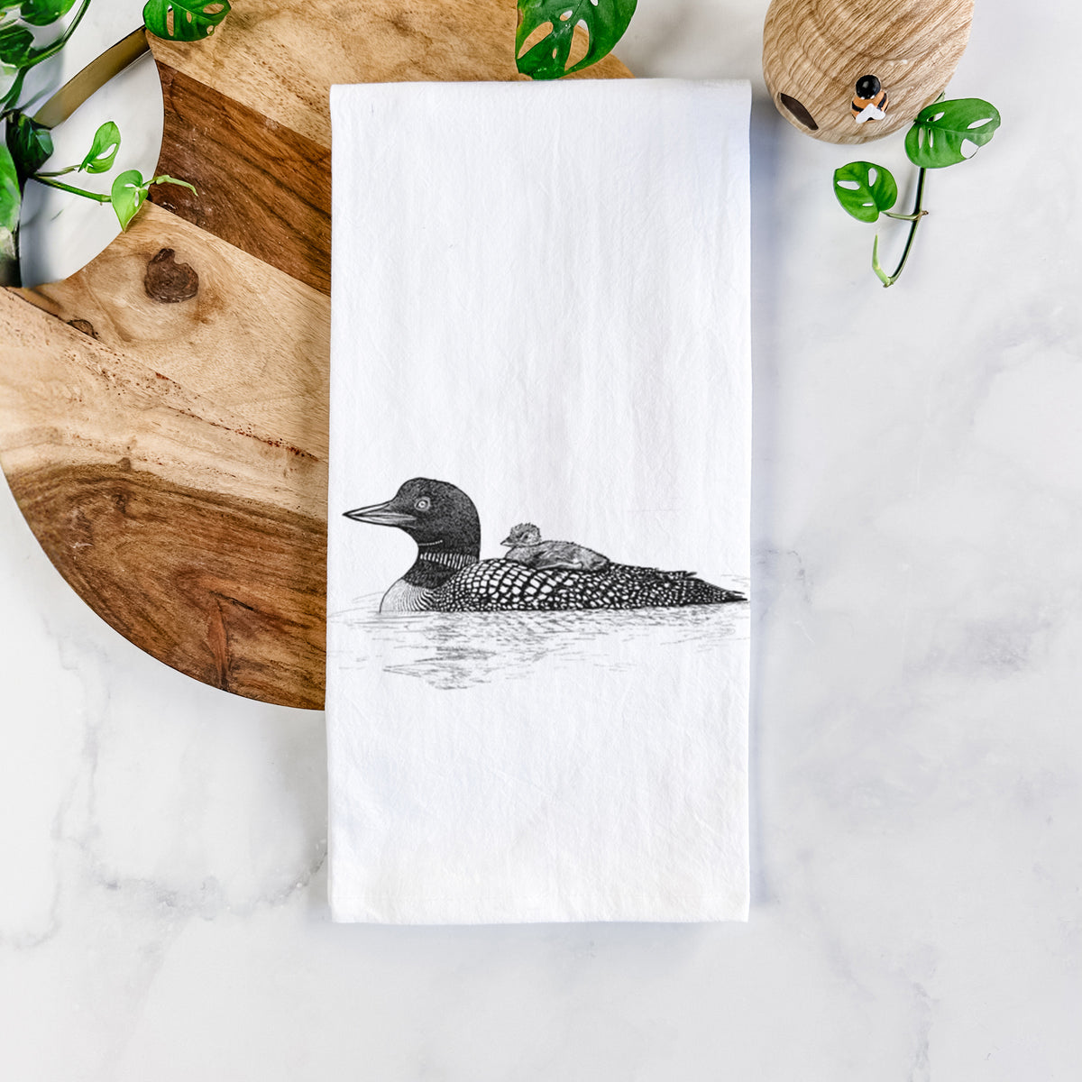 White cotton tea towel printed with a black ink sketch of a common loon floating on water with a chick riding on its back, on a marble counter beside a round wooden cutting board and greenery.