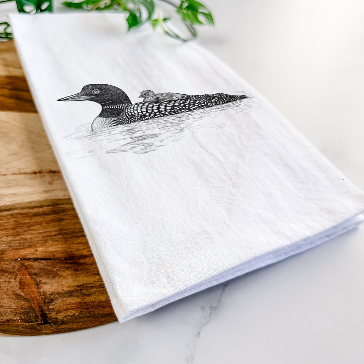 Close-up of a folded white tea towel on a wooden board, showing a black ink sketch of a common loon floating on water with a chick riding on its back.