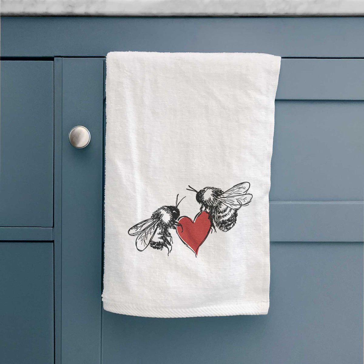 White hand towel printed with a black ink illustration of two bees facing each other with a red heart between them, hanging on a cabinet door.