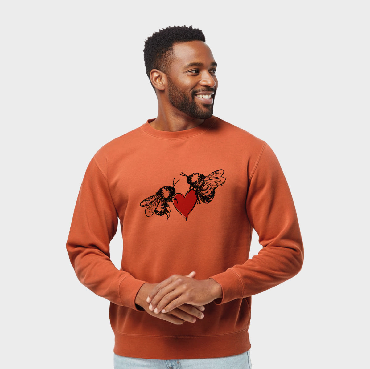 Model wearing an amber orange pigment dyed crewneck sweatshirt printed with a black ink illustration of two bees facing each other with a red heart between them.