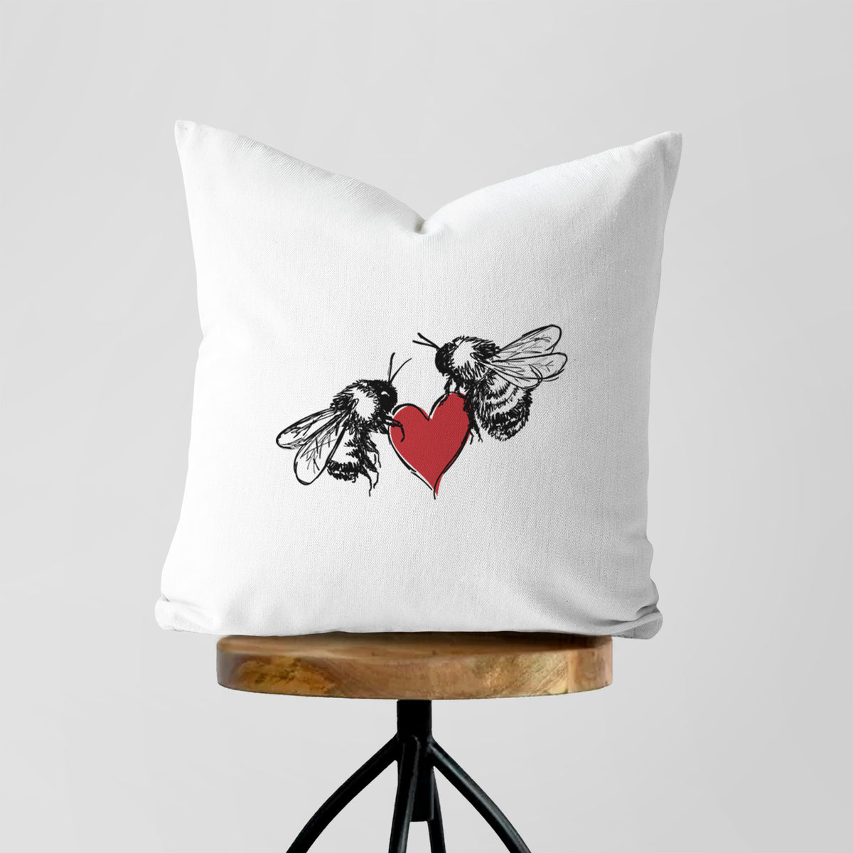 White pillow cover printed with a black ink illustration of two bees facing each other with a red heart between them, on a wooden stool.