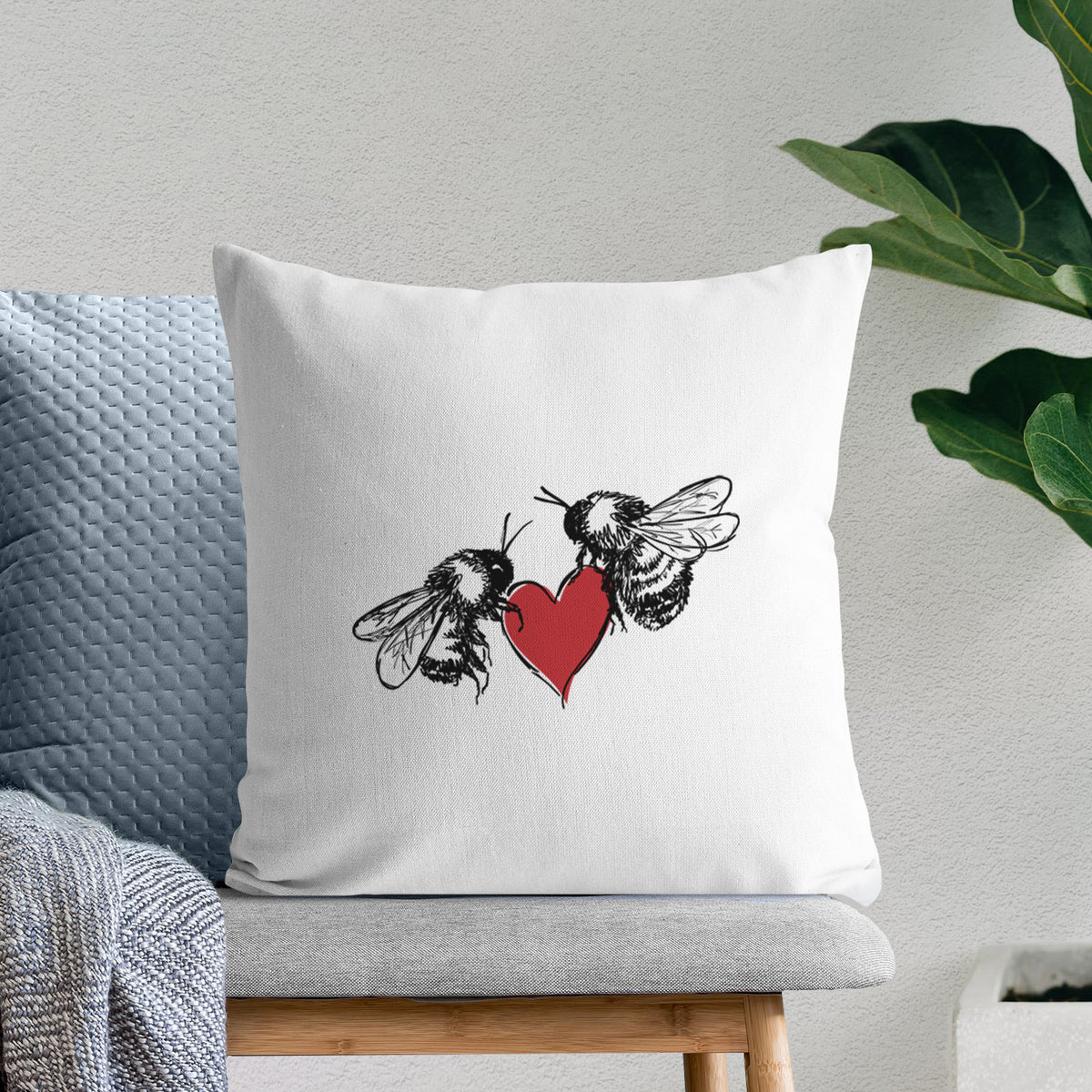 White pillow cover printed with a black ink illustration of two bees facing each other with a red heart between them, on a bench beside plants.