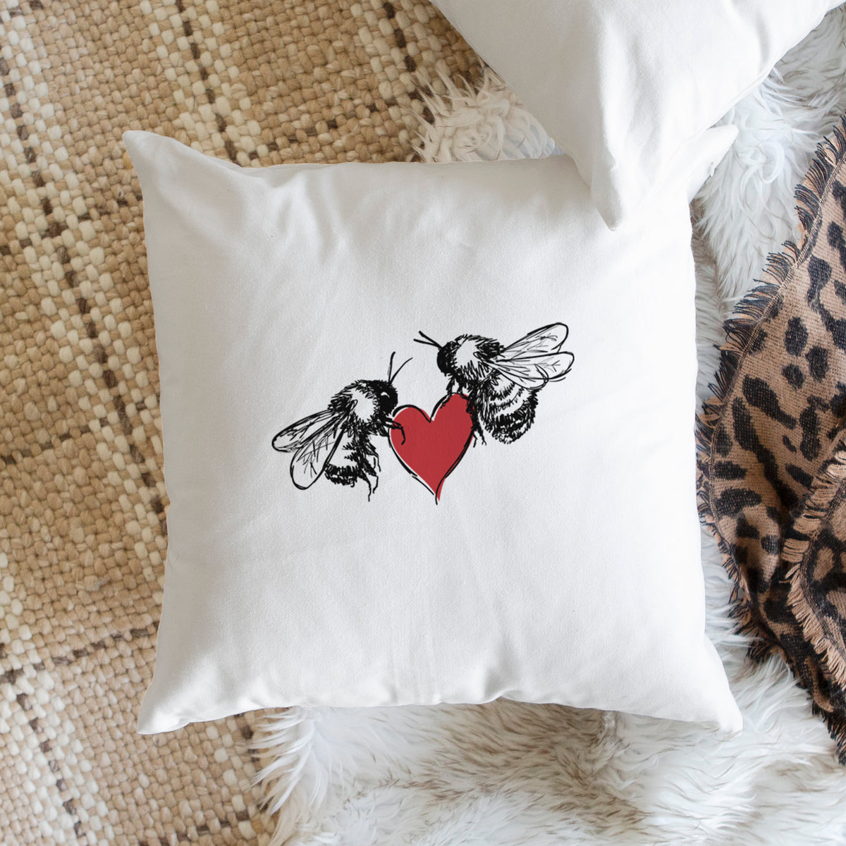 White pillow cover printed with a black ink illustration of two bees facing each other with a red heart between them, on a woven chair.