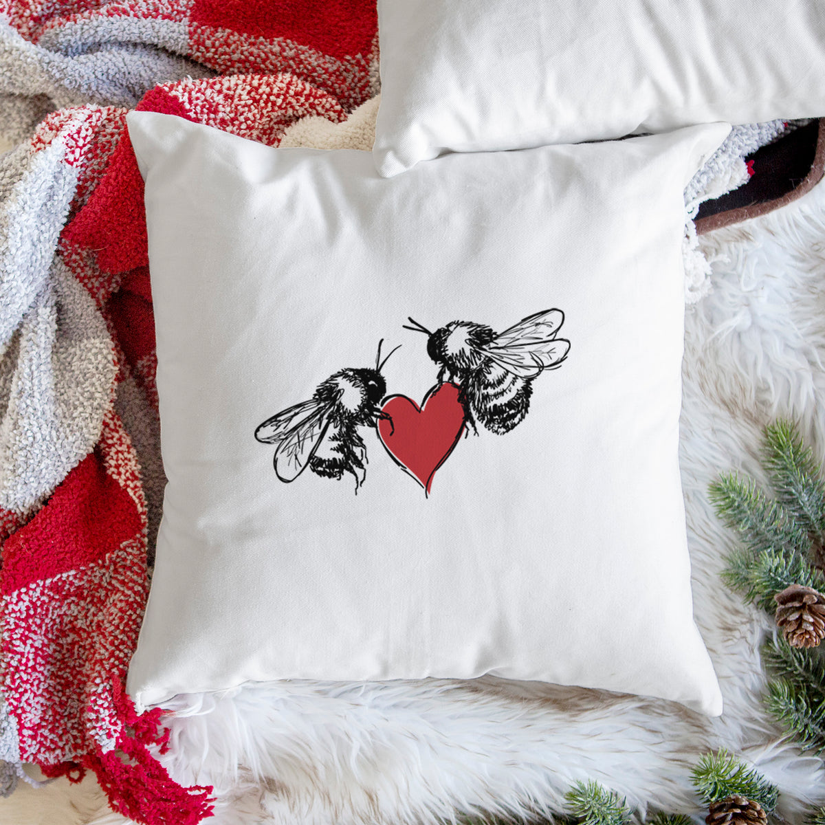 White pillow cover printed with a black ink illustration of two bees facing each other with a red heart between them, on a woven chair.