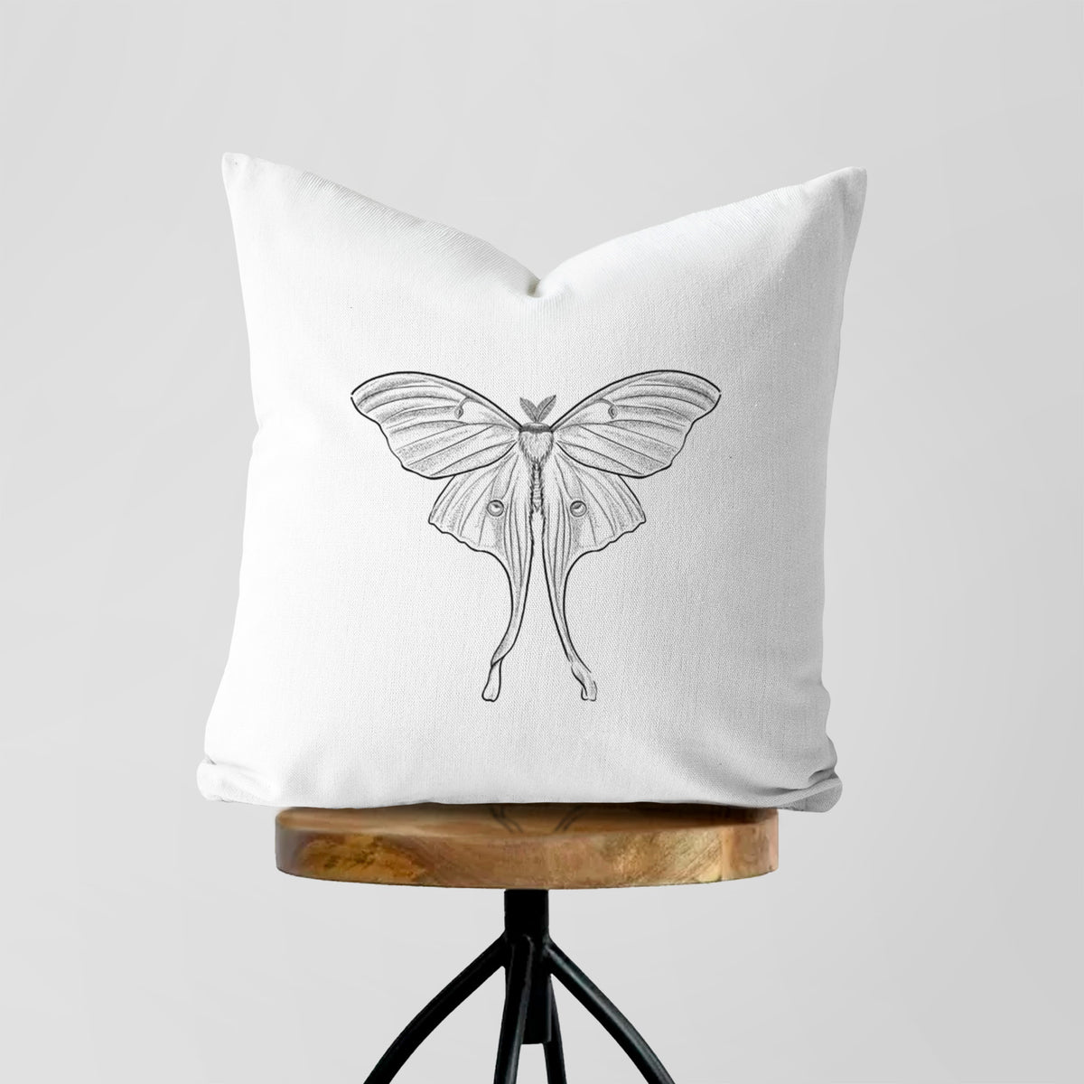 White pillow cover printed with a black ink line drawing of a luna moth with long trailing tails on its wings, on a wooden stool.