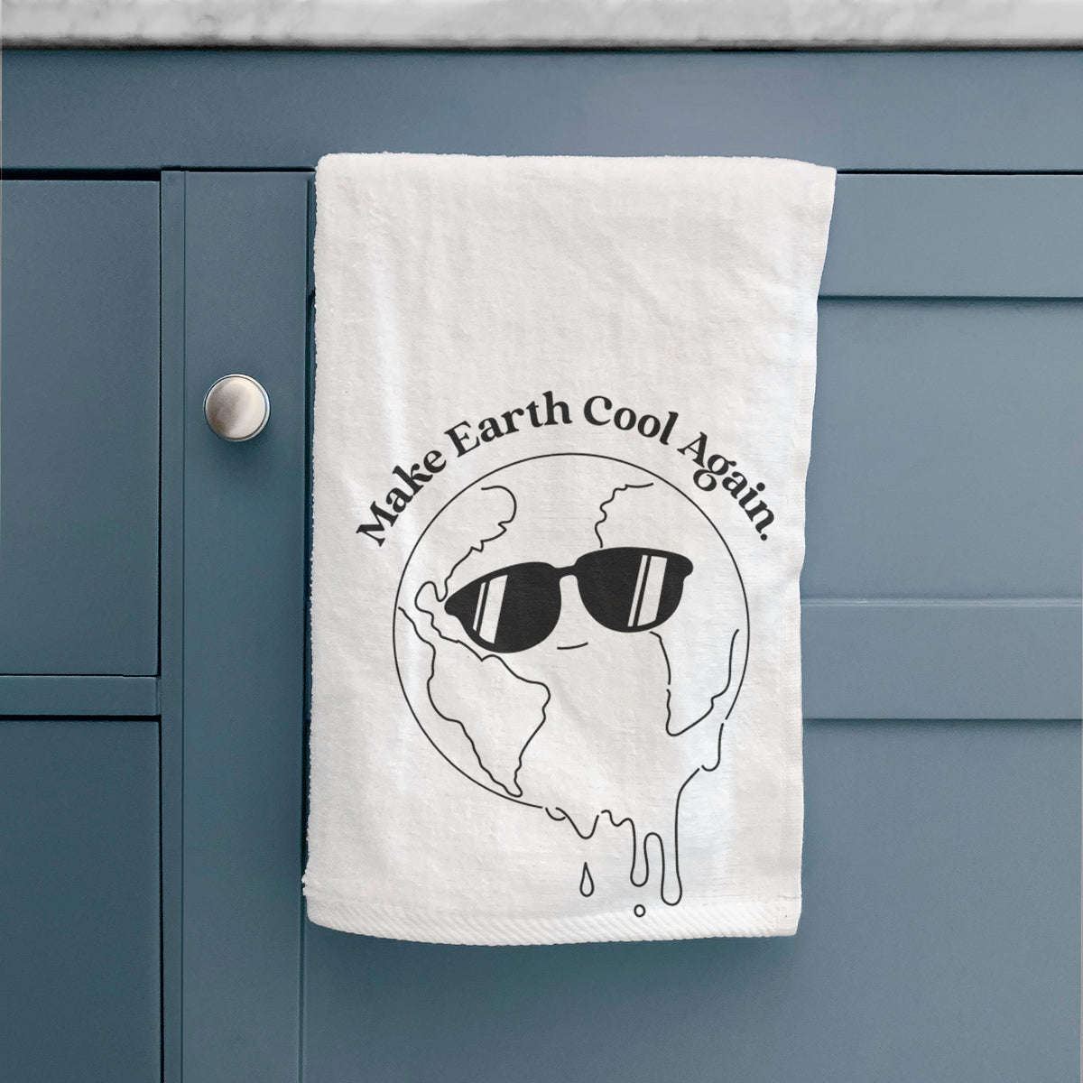 White hand towel printed with a melting Earth wearing black sunglasses, with the words Make Earth Cool Again, hanging on a cabinet door.