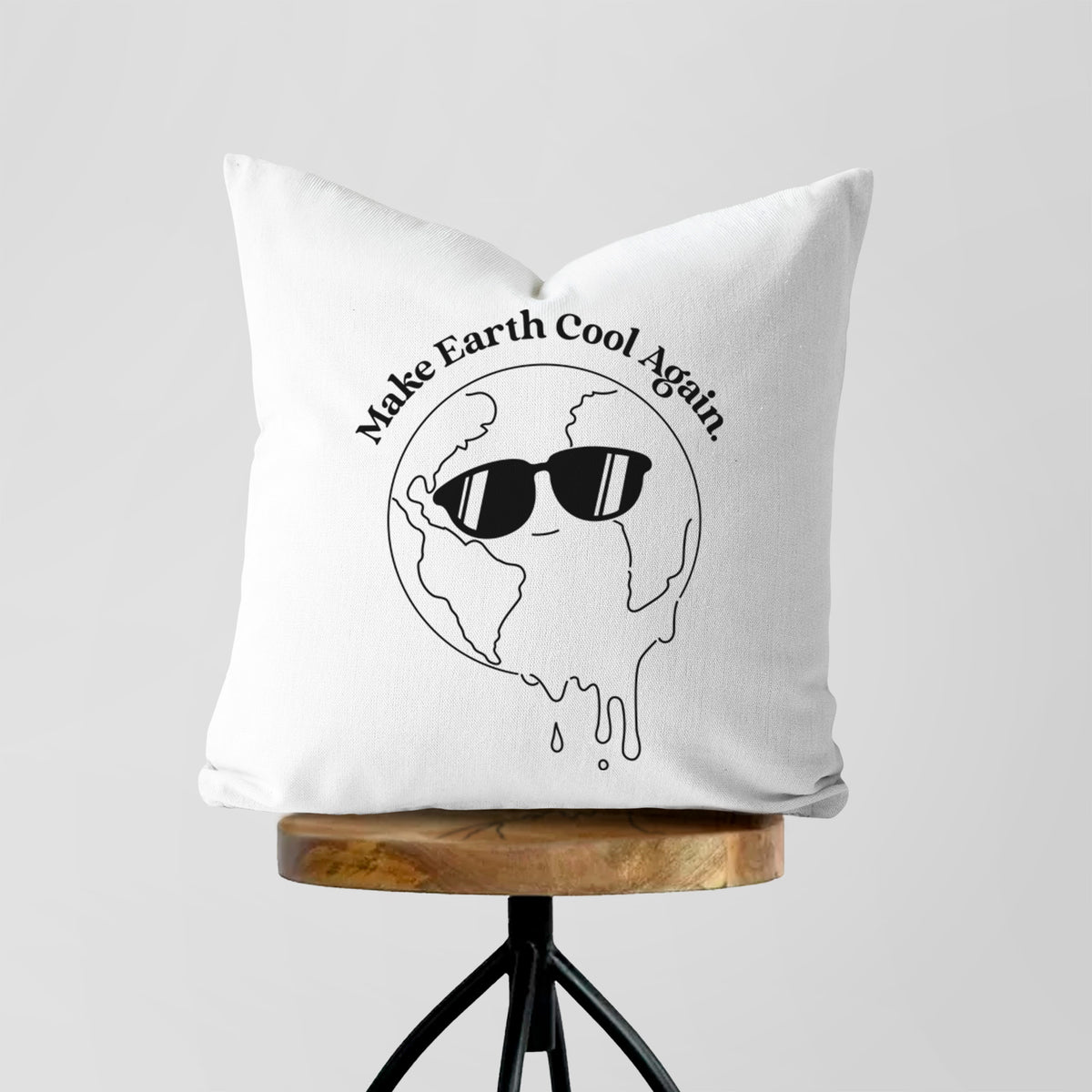 White pillow cover printed with a melting Earth wearing black sunglasses, with the words Make Earth Cool Again, on a wooden stool.