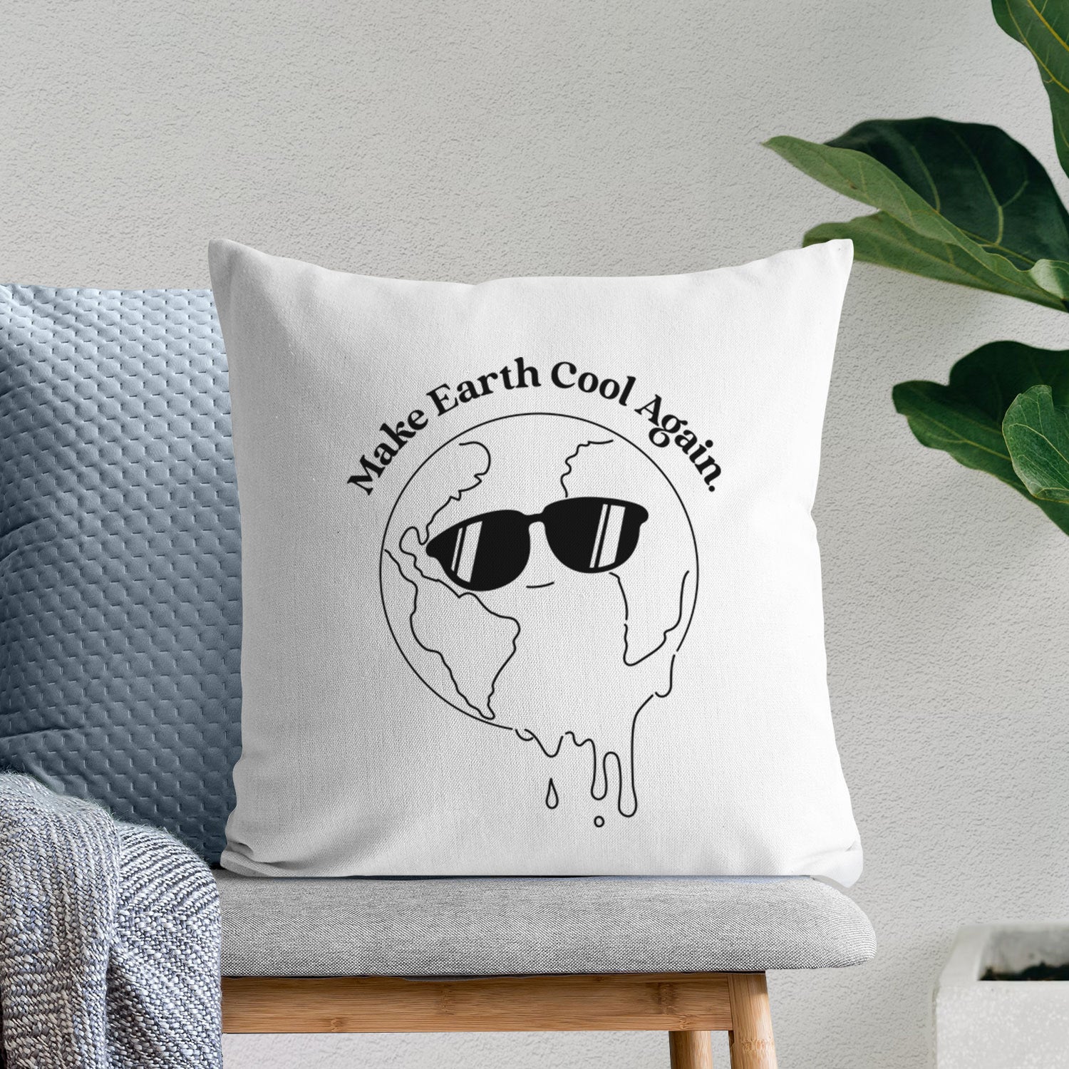 White pillow cover printed with a melting Earth wearing black sunglasses, with the words Make Earth Cool Again, on a bench beside plants.