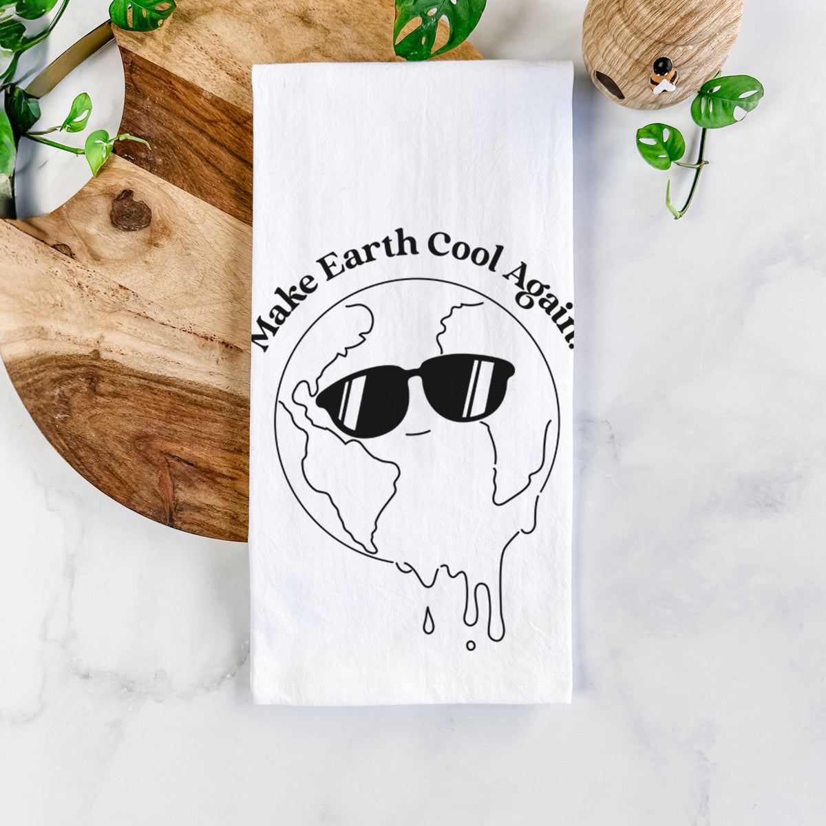 White cotton tea towel printed with a melting Earth wearing black sunglasses, with the words Make Earth Cool Again, on a marble counter beside a round wooden cutting board and greenery.