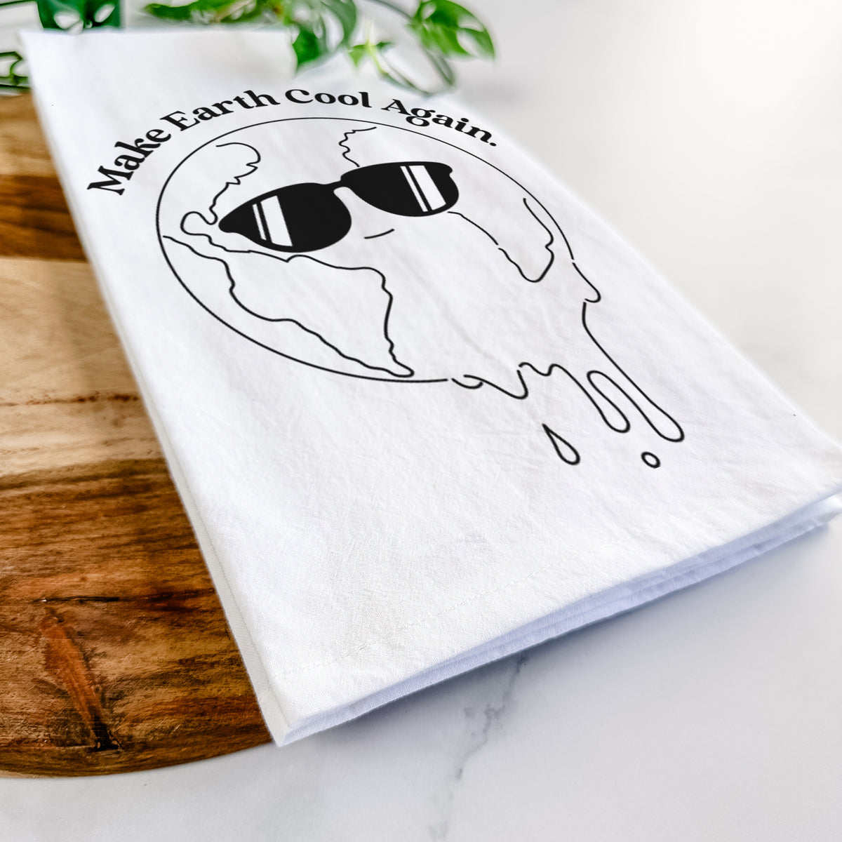 Close-up of a folded white tea towel on a wooden board, showing a melting Earth wearing black sunglasses, with the words Make Earth Cool Again.