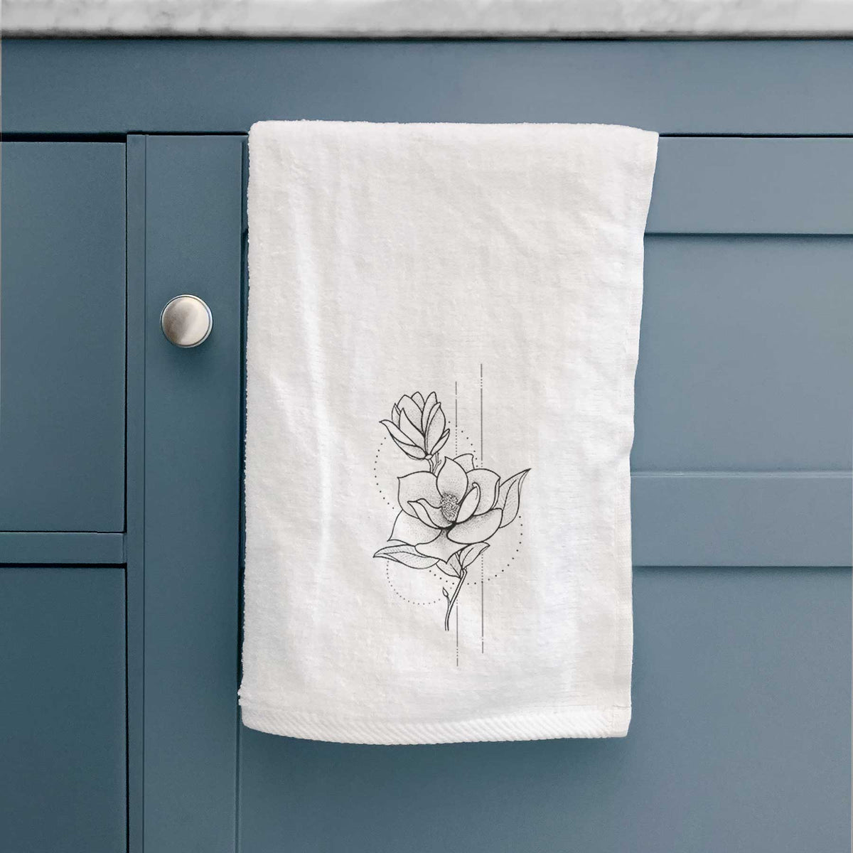 White hand towel printed with a black ink line drawing of a magnolia blossom on a stem, hanging on a cabinet door.